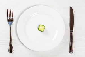 Eine Gurkenscheibe auf einem weißen Teller, neben Messer und Gabel - Konzeptbild Gewichtsverlust
