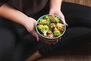 Eine Person im Schneidersitz hält eine Schüssel mit gesundem Salat mit Mikrogrün, Romanesco Kohl und Walnusskernen