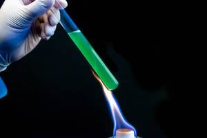 Eine Person mit Gummihandschuh erhitzt ein Reagenzglas mit grüner Flüssigkeit über einer Flamme mit schwarzem Hintergrund
