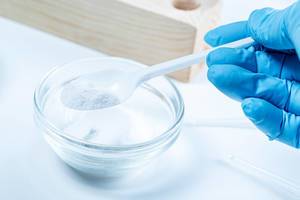 Eine Person mit Gummihandschuhen hält einen Plastiklöffel mit weißem Pulver über einer Glasschale in einem Labor
