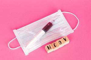 Eine Spritze mit Blutprobe liegt auf einer Gesichtsmaske neben der Bezeichnung NCoV auf Holzwürfeln auf pinkem Hintergrund