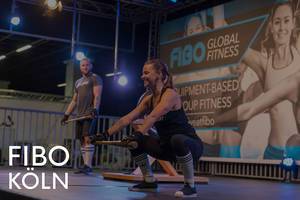Fitnesstrainerin benutzt Hilfsmittel für Sportübungen auf der Bühne, um ein Gruppen-Workout zu leiten, neben dem Bildtitel "Fibo Köln"