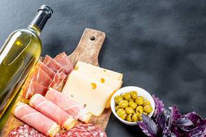 Flasche Weißwein, grüne Oliven und geschnittener Schinken und Käse auf schwarzem Hintergrund