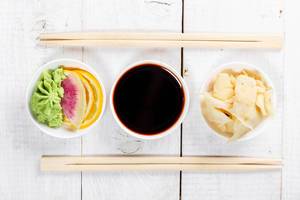 Flatlay von traditionellen Soßen für Sushi: Wasabi mit Zitrone, Sojasoße und Ingwer in weißen Schalen mit Essstabchen auf einem Holztisch