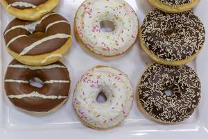 Flatlay zeigt drei verschiedene Donuts, mit Schokolade, bunten Streuseln und Zuckerglasur, auf einem weißen Tablett