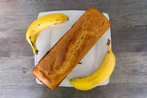 Flatlay zeigt selbstgebackenes Bananenbrot-Gebäck mit brauner Kruste, auf einem weißen Teller, auf einem Holztisch