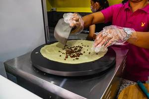 Frau gibt Schokostücke auf fast fertigen süßen Pfannkuchen auf spezieller Crêpe-Kochplatte