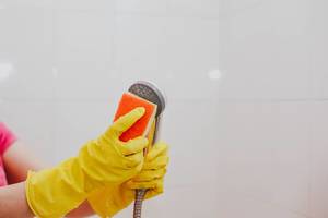 Frau mit gelben Gummihandschuhen und einem Schwamm reinigt die Duschbrause im Badezimmer am Putztag