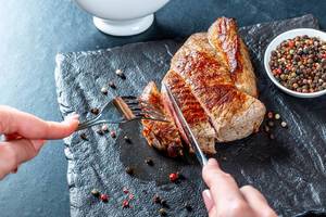 Frau schneidet frisches Steak auf Schieferplatte, daneben verschieden farbige Pfefferkörner