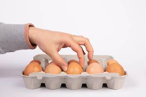 Frauenhand nimmt braunes Hühnerei aus gefülltem Eierkarton von weißem Hintergrund