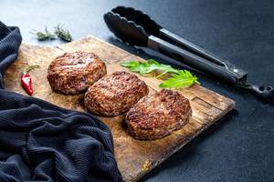 Frisch gegrilltes Burgerfleisch auf einem alten rustikalen Holzbrett, auf schwarzem Hintergrund