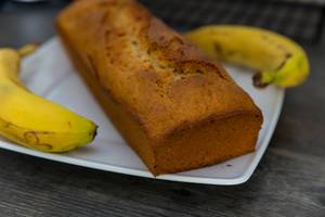 Frischgebackenes Bananenbrot, mit knuspriger Kruste, auf einem weißen Teller, neben zwei Bananen