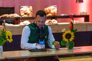 Frühstückspause auf der Bits & Pretzels Messe: Mann in bayrischer Tracht sitzt mit Brötchen an der Holzbar und nutzt das Tablet