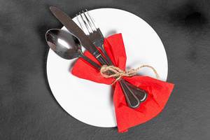 Gebundenes Gedeck aus Löffel, Messer, Gabel und roter Serviette auf weißem Teller und schwarzem Hintergrund