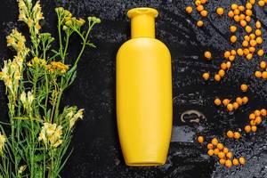 Gelbe Kunststoffflasche, Sanddornbeeren und Wildblumen auf schwarzem Hintergrund mit Wassertropfen - Naturkosmetik