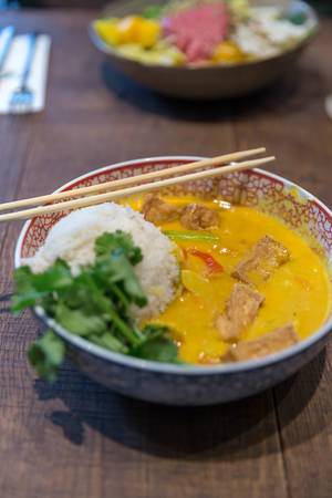 Gelbes Kokos-Tofu-Curry als asiatisches veganes Mittagessen mit Duftreis, Blumenkohl & Babyspinat, in einer Schüssel mit Essstäbchen, auf einem Holztisch