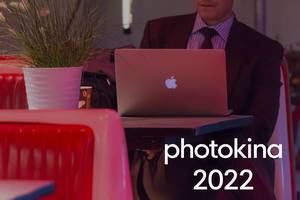Geschäftsmann sitzt mit einem Kaffee-to-go-Becher im Café vor seinem Macbook, neben dem Bildtitel "Photokina 2022"