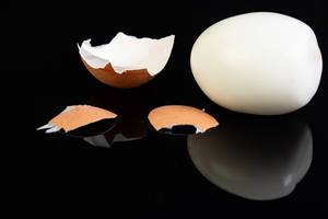Geschältes hartgekochtes Ei neben Eierschale auf spiegelndem schwarzem Hintergrund