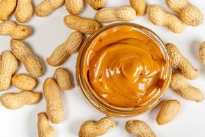 Glass jar of peanut butter with peanuts on white background, top view