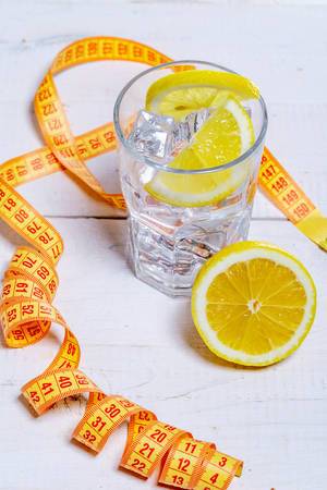 Glass of cold water with ice cubes and lemon on white wooden background with measuring tape-healthy lifestyle concept