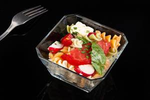 Glasschale mit Nudelsalat mit Tomatenscheibe, Rucola, Radieschen und Fetakäse vor schwarz