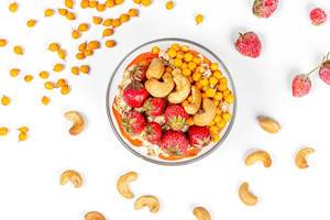 Glasschüssel mit Haferflocken, Cashew Nüssen, Erdbeeren und Sanddornbeeren mit Zutaten auf weißem Hintergrund verstreut von oben fotografiert