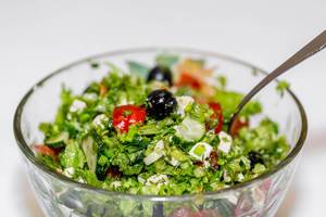 Griechischer Salat mit Oliven, Tomaten, Fetakäse und Kräutern in Glasschale vor weißem Hintergrund