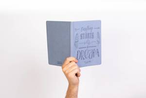 Hand hält ein blaues Notizbuch nach oben, mit dem Covertext: Alles beginnt mit einem Traum