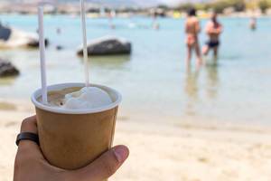 Hand hält einen kalten, griechischen Frappé mit Eiskugel, am Strand von Paros