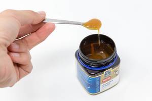 Hand hält einen Löffel mit flüssigem Honig über da Honigglas "Manuka Honey"