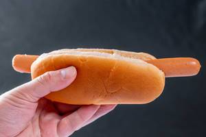 Hand hält Hotdog bestehend nur aus Wurst und Brötchen vor dunklem Hintergrund