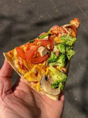 Hand hält veganes Pizzastück Kap Verde von Dominos als Streetfood mit Tomate, Brokkoli, Pilzen mit veganem Käse überbacken in Köln