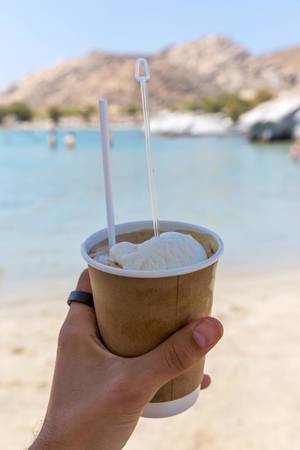 Hand hold a cold Greek frappé coffee with ice cream scoop, in front of the blue sea at the beach of Paros
