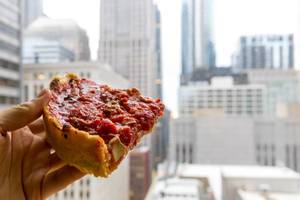 Hand holding a slice of Chicago-style pizza with cheese and tomato made by Pizzeria Uno, creators of the first deep dish pizza in 1943. In the background, the skyscrapers of Downtown Chicago