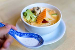 Hand holding an Asian rice spoon, in front of vegan starter "Tom Yum", coconut milk soup with healthy vegetables & tofu as meat substitute