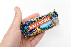Hand holding "Hafervoll Flapjack Mexican" - cereal bar with colorful packaging showing two maracas in front of a white background