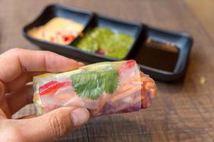 Hand holds a summer roll filled with avocado, mango, paprika, red cabbage, carrots and rice noodles with various sauces in the background