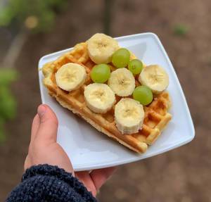 Hand holds a waffle with banana slices and green grapes on a white plate