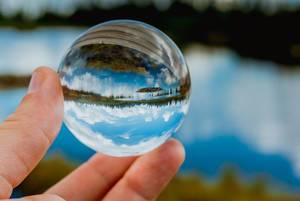 Hand holds glass ball showing landscape with lake, woodland and blue sky with clouds