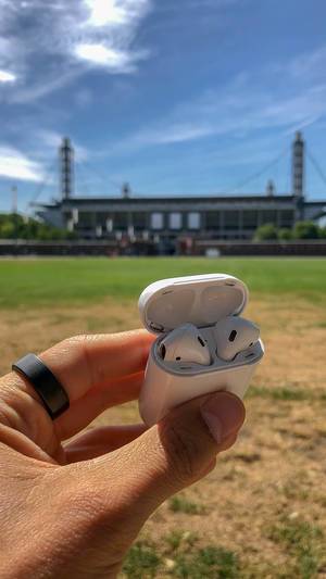 Hand holds white iPhone AirPods headphones in front of the RheinEnergie Stadium in Cologne