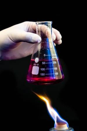 Hand in glove holds flask with chemical solution over fire on black background.