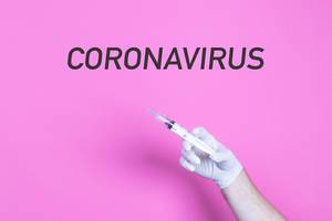 Hand mit medizinischen Handschuhen hält eine Spritze unter den Worten Coronavirus