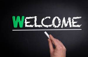 Hand schreibt das Wort "Welcome" - Willkommen - mit Kreide auf eine schwarze Tafel
