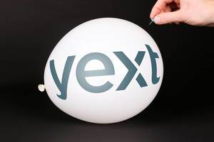 Hand uses a needle to burst a balloon with Yext logo