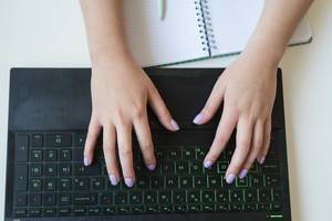 Hands of a woman typing on a black laptop keyboard and a squared notebook with pen seen from above