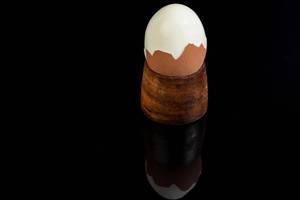 Hartgekochtes Ei halb geschält in hölzernem Eierbecher vor reflektierendem schwarzem Hintergrund