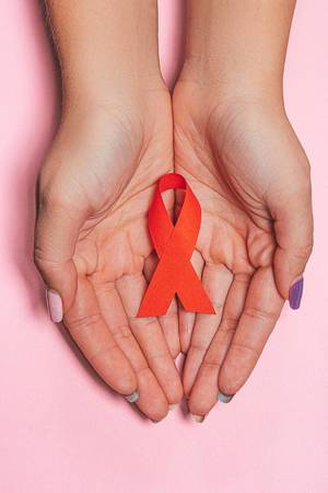 Healthcare and medicine concept - female hands with red AIDS awareness ribbon on pink background