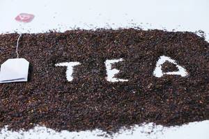 Hintergrundbild zum Thema Tee: Das Wort "Tea" in getrocknetem Tee, neben einem Teebeutel auf einem weißem Tisch