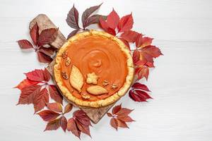 Homemade pumpkin pie with red autumn leaves on white wooden table. Top view