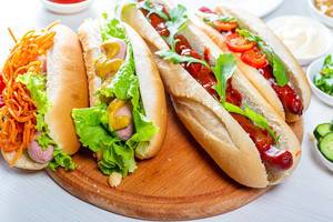 Hotdogs mit verschiedenen Würstchen und bunten Zutaten, wie Karotten, Salat, Tomaten, Senf und Ketchup, auf einem runden Holzbrettchen